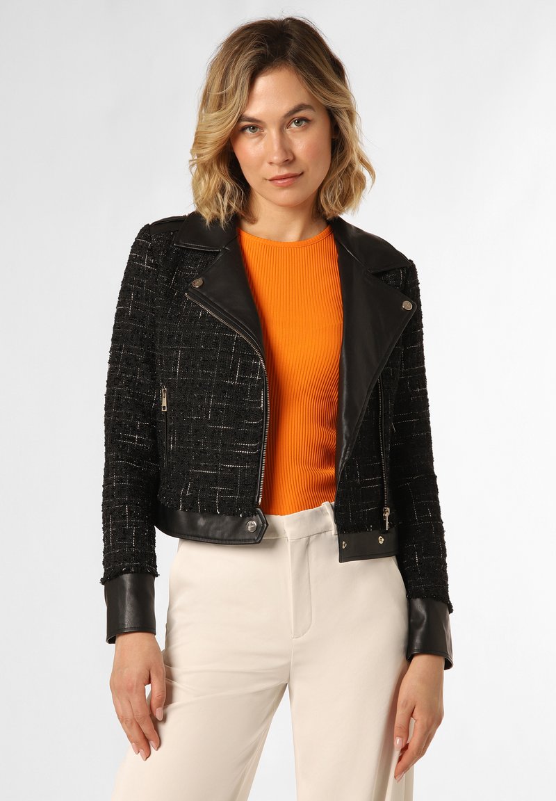Schwarze, strukturierte, kurz geschnittene Jacke mit Lederakzenten, silbernen Reißverschlüssen und Druckknopfverschluss. Kombiniert mit einem leuchtend orangefarbenen gerippten Oberteil.