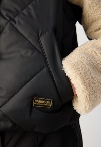 Μαύρο καπιτονέ μπουφάν με αφράτο μπεζ γιακά και μανσέτες. Διαθέτει χρυσή ετικέτα "Barbour International" στο μπροστινό μέρος.