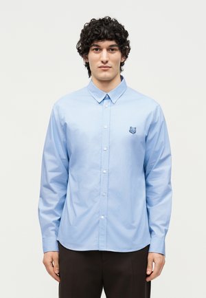 Mann mit lockigem Haar, der ein hellblaues, geknöpftes Hemd mit einem kleinen bestickten Bärenlogo auf der linken Brust und dunkle Hosen trägt.