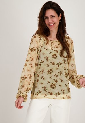 Vrouw glimlachend, gekleed in een doorschijnende beige blouse met een bruin bloemmotief en witte broek, tegen een effen lichte achtergrond.