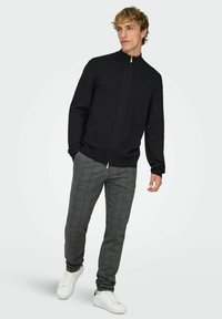 Maglione nero con zip e collo alto, abbinato a pantaloni a quadri grigi e sneakers bianche. Tessuto liscio, design aderente, stile moderno.