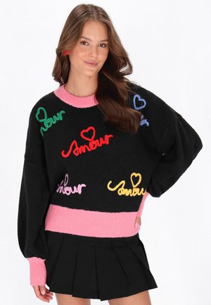 Jonge vrouw met golvend bruin haar, gekleed in een zwarte trui met roze boorden en Franse woorden "amour" in kleurrijk garen, gecombineerd met een zwarte plissérok.