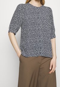 Námořnická halenka s bílým květinovým vzorem, krátkými nabíranými rukávy, knoflíkovým zapínáním a měkkou tkaninou. Spárovaná s hnědými kalhotami s širokými nohavicemi.