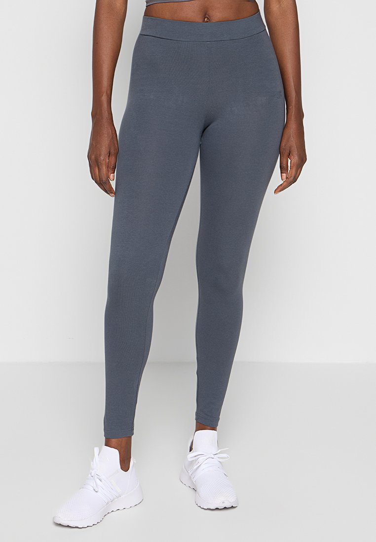 Leggings gris extensibles avec une taille haute, une texture lisse et une coupe ajustée. Portés avec des chaussures de sport blanches.