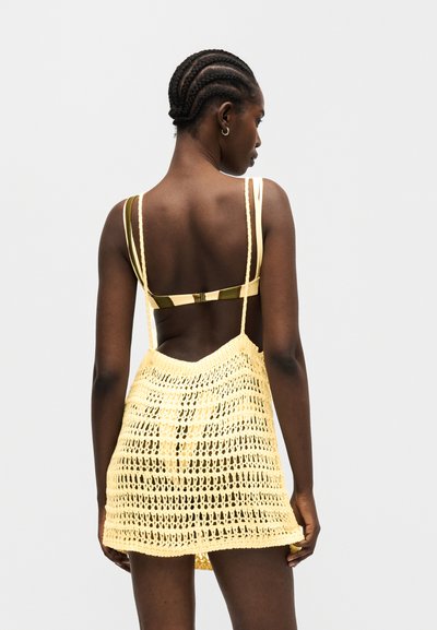 Γυναίκα με πλεξούδες φοράει κίτρινο και πράσινο ριγέ μπικίνι και κίτρινη πλεκτή παραλία φούστα, με την πλάτη γυρισμένη σε απλό φόντο.