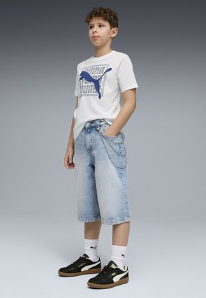 Junge steht mit den Händen in den Taschen, trägt ein weißes Puma-T-Shirt, helle Jeansshorts mit Ketten, weiße Puma-Socken und schwarze Puma-Sneaker.
