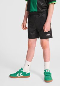 Svarta idrottsshorts med randig design och "hummel"-logotyp, tillsammans med gröna sneakers med vita detaljer och kardborreband, samt vita strumpor.