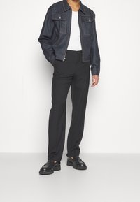 Hombre vestido con una chaqueta de mezclilla oscura sobre una camisa blanca, pantalones de vestir negros y mocasines de cuero negros, de pie contra un fondo blanco.