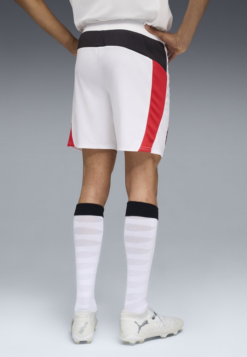 Shorts blancs avec des accents noirs et rouges, chaussettes blanches texturées avec des poignets noirs, et crampons de football argentés. La vue arrière montre une coupe athlétique.