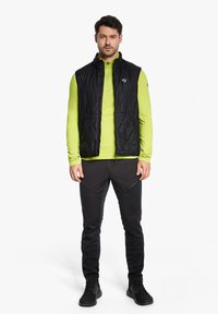 Black puffer vest with zippered pockets over a lime green long-sleeve shirt, paired with dark tapered pants and black athletic shoes.
