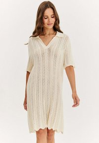 Sukienka dzianinowa w kolorze kremowym z dekoltem w serek, krótkimi rękawami i falbanym wykończeniem. Materiał ma teksturowany, ażurowy wzór z pionowymi paskami.