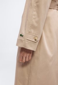 Μανσέτα μπεζ παλτομανικιού με ραμμένη λουρίδα και μικρό πράσινο λογότυπο κροκόδειλου πάνω από τον καρπό, χέρι χαλαρό δίπλα στο σώμα.