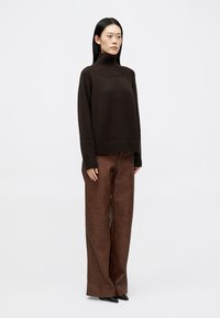 Maglione a collo alto marrone in maglia con dettagli a coste abbinato a pantaloni ampi in finta pelle marrone. Il modello si trova in posizione eretta, con le mani ai lati.