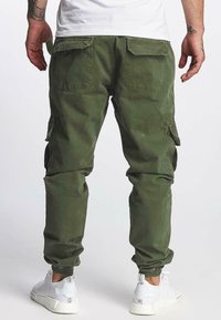 Olijfgroene cargobroek met elastische boorden en zijzakken, gemaakt van een duurzaam materiaal. Draagt met witte sportieve schoenen.