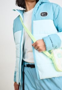 Ljusblå pufferjacka med huva, utrustad med en svart "AIR"-logotyp och detalj av "THERMA-FIT". Kombinerad med en pastellgrön axelväska.