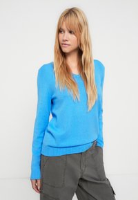 Ανοιχτό μπλε πουλόβερ με V-λαιμόκοψη και ριμπ μανσέτες. Μαλακή υφή, ελαφρώς εφαρμοστή σιλουέτα, συνδυασμένο με σκούρο παντελόνι cargo.