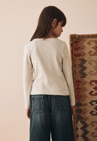 Chemise beige côtelée à manches longues avec un col rond, associée à un jean denim bleu à jambes larges avec des poches arrière. Vue de dos.