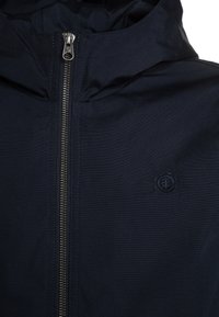 Giacca a vento blu navy con zip frontale, colletto alto e un piccolo logo sul lato sinistro. Tessuto liscio con un design aderente.
