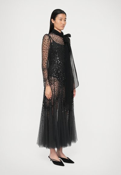 Naine kannab läikivat musta kleiti, mis on kaunistatud sädeluste ja sametpunktidega, koos mustade terava ninaga slingback kingadega, seistes valge taustaga.