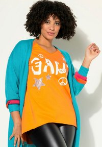 Turkis lang cardigan over en orange V-hals t-shirt med sølv stjerner og fredssymboler, parret med sorte leggings. Glatte teksturer.