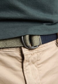 Woven fabric belt w kolorze oliwkowym i granatowym, z okrągłą metalową klamrą. Beżowe spodnie o subtelnej teksturze są częściowo widoczne.