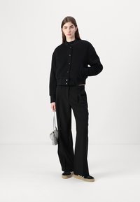 Giacca bomber nera cropped con bottoni, abbinata a pantaloni larghi neri, sneakers neri e una piccola borsa metallizzata.