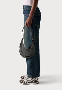 Black and white studded handbag with a curved design, short chain strap, held in a hand alongside blue jeans and brown and white sneakers.