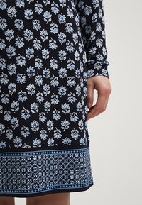 Robe bleu marine avec motif floral bleu clair, bordure géométrique près de l'ourlet, manches longues, et une main reposant sur le côté.