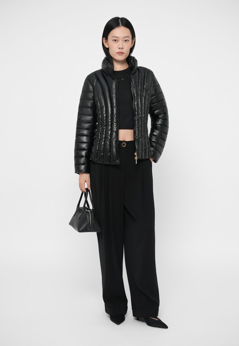 Veste matelassée noire à design vertical, associée à un crop top noir et un pantalon à jambes larges. Un petit sac à main noir complète la tenue.