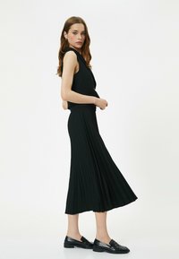 Zwarte geplooide jurk met een getailleerde corsage en een vloeiende rok, gecombineerd met zwarte patenten instappers. Soepele textuur en elegante silhouette.