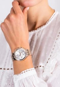 Reloj de metal plateado con cara redonda, tres subdiales y un bisel acentuado con cristales, usado en una muñeca sobre un fondo de tela clara.