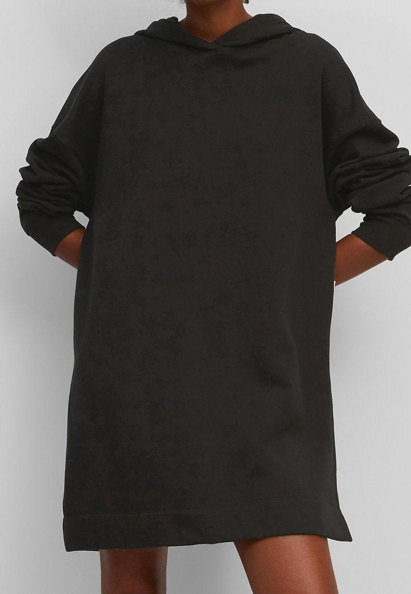 Personne portant une robe sweat à capuche noire ample avec manches longues et capuche, mains reposées dans le dos.