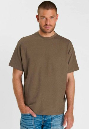 Man met kort haar en blauwe ogen die een eenvoudige bruine korte mouwen shirt en een blauwe spijkerbroek draagt, staand met één hand in de zak.