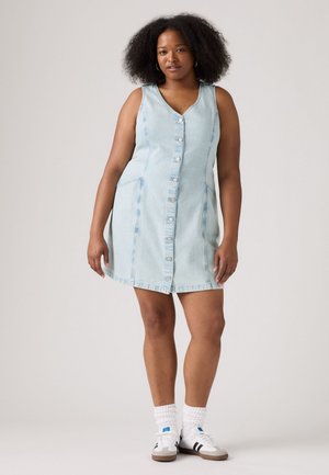 Light blue denim sleeveless dress with front buttons, side pockets, and a slightly flared shape. Worn with white textured socks and sneakers.