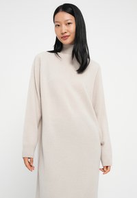 Jonge vrouw met stijl zwart haar die een lange, beige coltrui jurk draagt tegen een effen witte achtergrond.