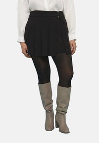 Falda mini negra con pliegues y detalle de cinturón, combinada con medias negras transparentes y botas hasta la rodilla en tono beige. Textura suave, diseño ajustado.