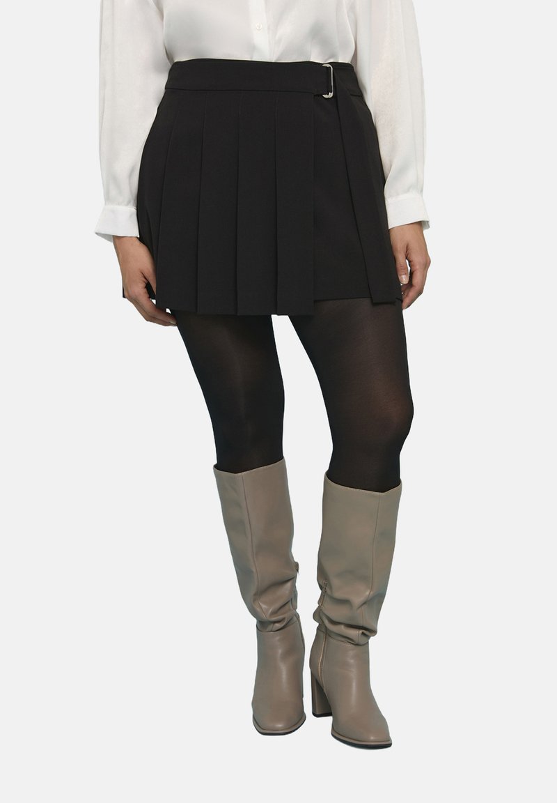 Falda mini negra con pliegues y detalle de cinturón, combinada con medias negras transparentes y botas hasta la rodilla en tono beige. Textura suave, diseño ajustado.