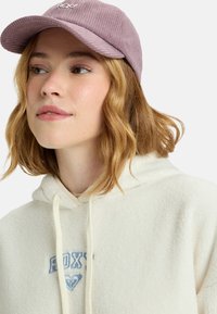 Paarse corduroyen pet met een gebogen rand; gecombineerd met een crèmekleurige fleece hoodie met blauwe "ROXY" borduring en een trekkoord.