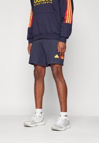 Námořnická mikina Adidas s žlutým logem a červenými pruhy, spárovaná s námořnickými kraťasy s žlutým trojúhelníkovým logem. Šedé sportovní boty s bílými ponožkami.