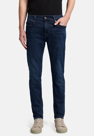 Dunkelblaue Denim-Jeans mit schmalem Schnitt, Fünf-Taschen-Design und Knopfverschluss. Tragen mit beigen Sneakers und einem schwarzen T-Shirt.