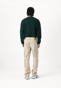 Un pull en tricot vert et duveteux associé à un pantalon cargo beige avec des poches latérales. Le modèle se tient de dos, mettant en valeur les textures du vêtement.