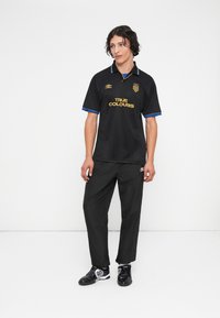 Νεαρός άντρας με σγουρά μαλλιά, φορώντας μαύρη αθλητική μπλούζα με χρυσό κείμενο "TRUE COLOURS" και μαύρο παντελόνι, στέκεται απέναντι σε βάθος λευκού.