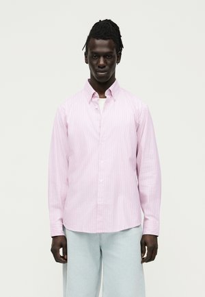 Άνδρας με σκούρο δέρμα και πλεξίδες, φοράει ένα ανοιχτό ροζ ριγέ πουκάμισο με κουμπιά και ανοιχτά μπλε παντελόνια, στέκεται μπροστά σε μία μονόχρωμη επιφάνεια.