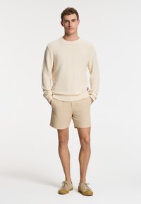 Beige knit sweater with ribbed cuffs, paired with light beige shorts and tan shoes. The outfit has a casual, relaxed fit.