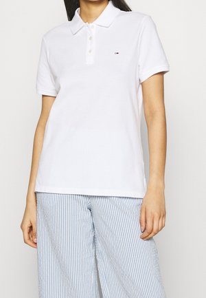 Frau trägt ein weißes kurzärmeliges Poloshirt mit kleinem Logo auf der Brust und hellblaue gestreifte Hosen mit weitem Bein.