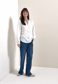 Blouse en satin blanc avec une patte de boutonnage et des manches longues, associée à un pantalon évasé en denim bleu et des sandales grises sur un fond neutre.
