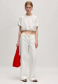 Witte cropped top met korte mouwen, hoog aangesloten witte broek en zwarte sandalen. Houdt een rode tas vast. Eenvoudig ontwerp, comfortabele pasvorm.