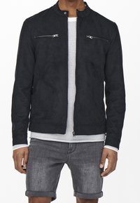 Giacca in suede nera con colletto alto, dotata di tasche sul petto con zip e una texture liscia. Indossata sopra una camicia bianca a fantasia e pantaloni corti grigi.