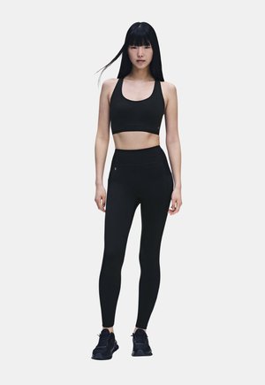 Jeune femme debout portant un soutien-gorge de sport noir, un legging noir taille haute et des chaussures de sport noires sur fond blanc.
