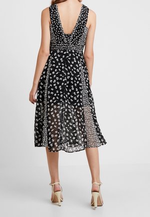 Femme portant une robe midi noire sans manches avec des motifs floraux blancs et une jupe inférieure transparente, associée à des sandales à talons hauts beiges.